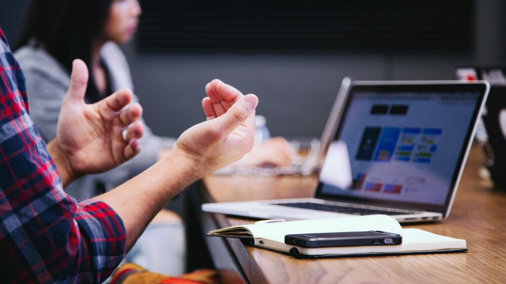 Someone uses their hands as they talk. There is a laptop on the desk in front of them beside a notebook and mobile phone.